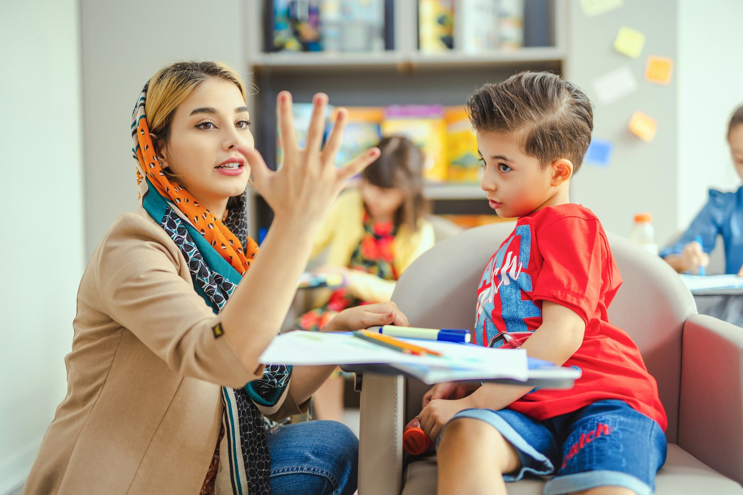 An adult holding up an open hand as she talks with a young boy seated across a low table of markers and paper