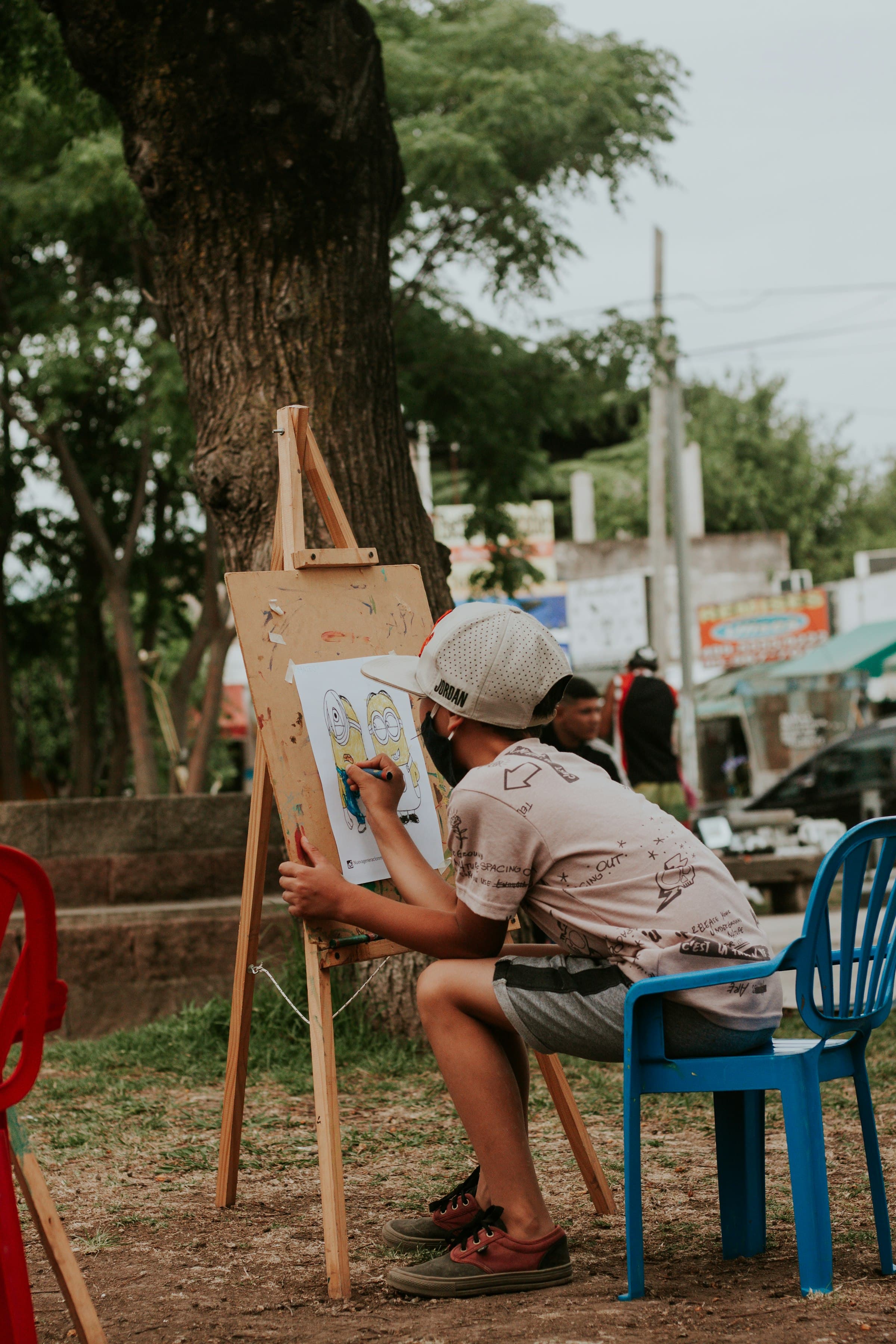 A child seated at a wooden easel outdoors under a tree, drawing on a sheet of paper