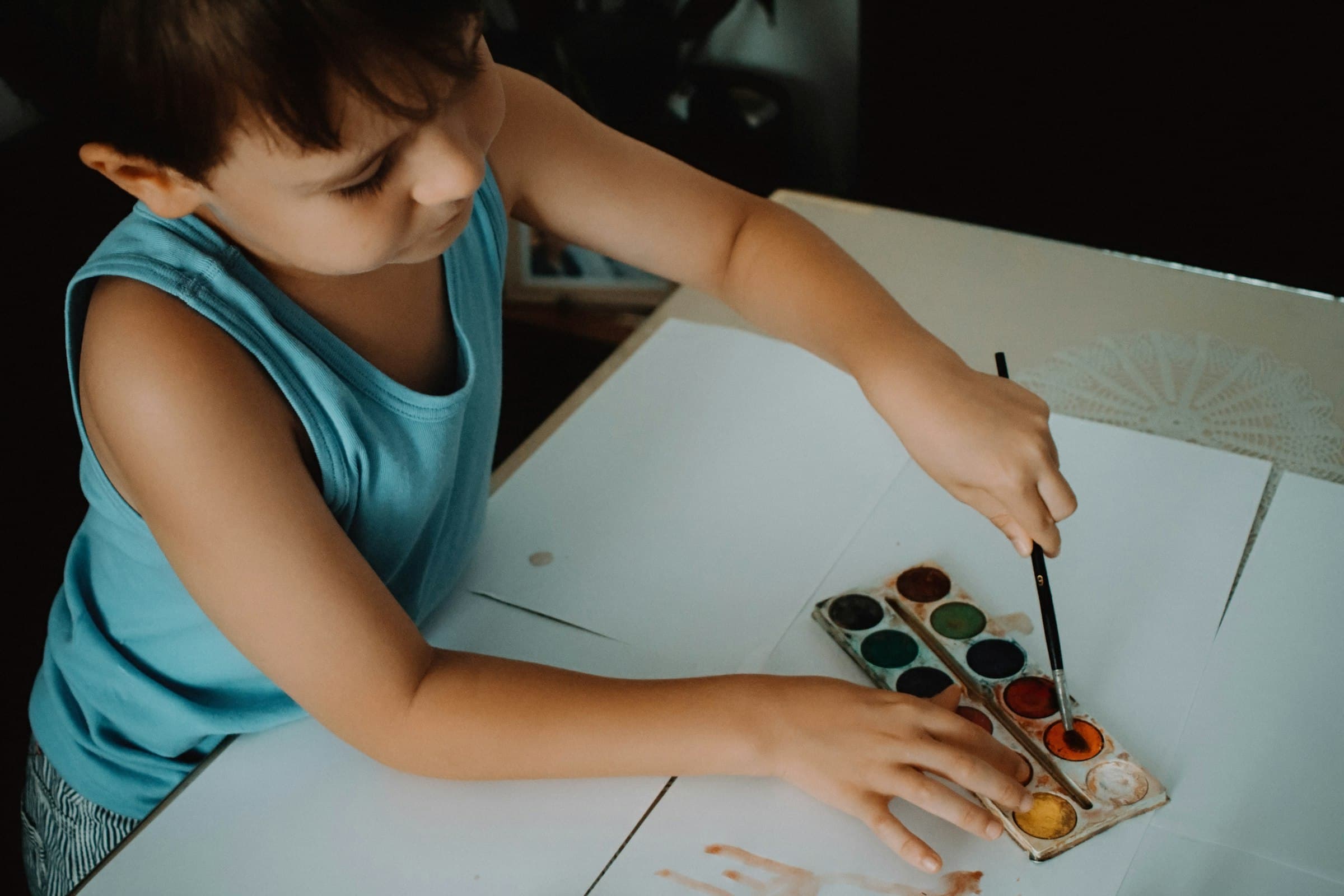 A young child at a table loading a brush from a tray of watercolour paints
