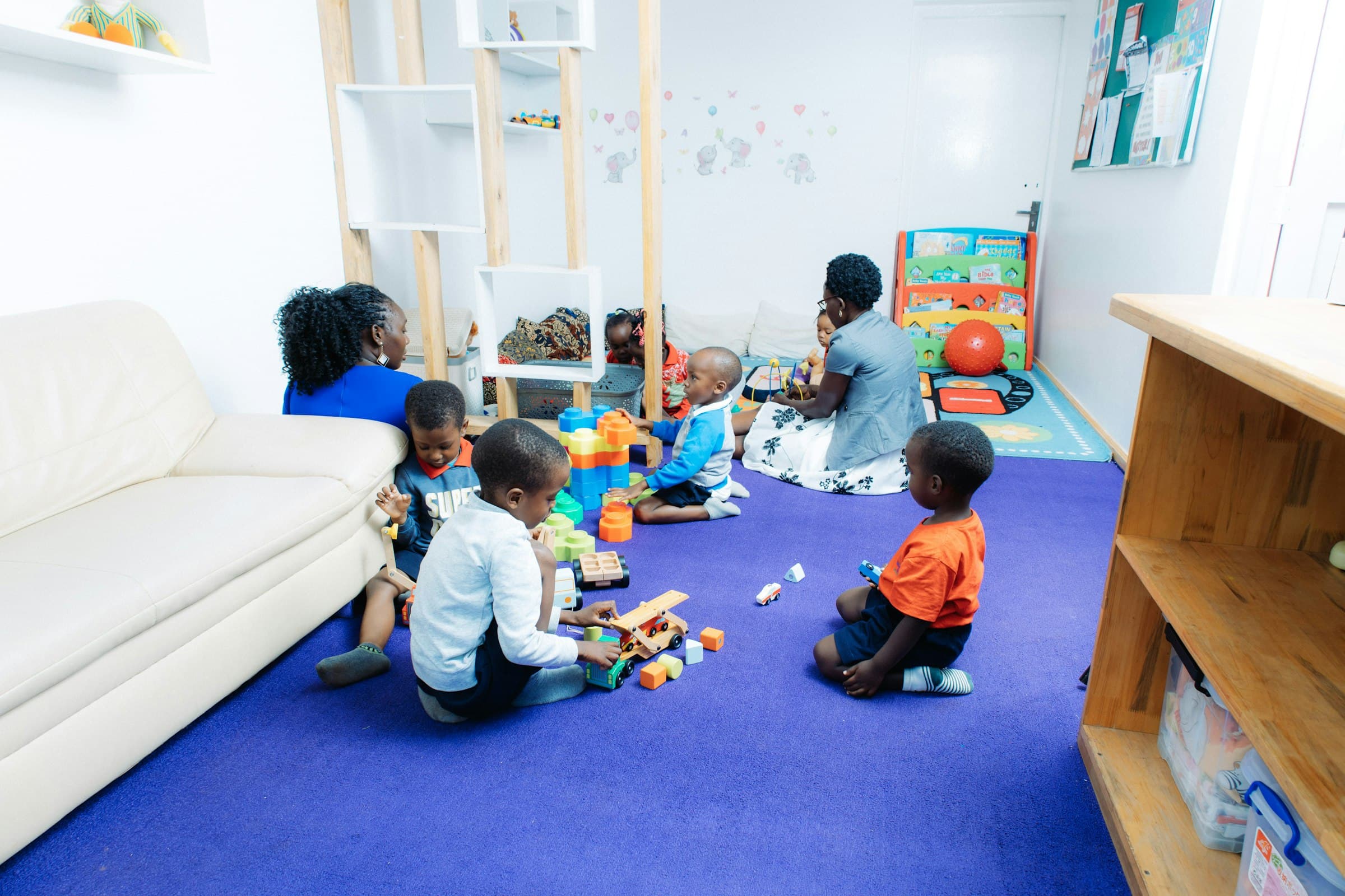 Young children playing with blocks and wooden toys on a bright playroom floor while two adults sit nearby