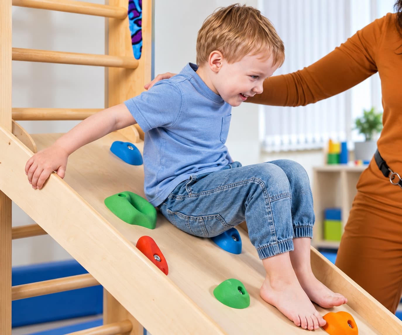 A young boy sliding down a wooden climbing ramp fitted with colorful holds, an adult's hand on his shoulder