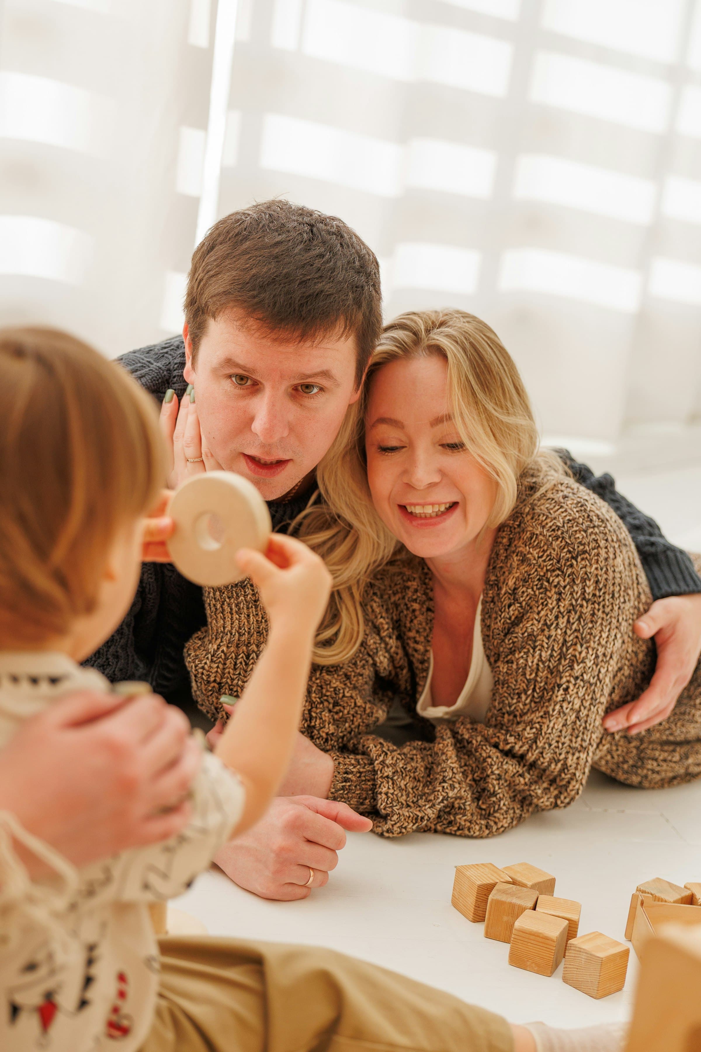 Two adults lying on the floor smiling as a young child holds up a wooden ring beside a scatter of wooden blocks